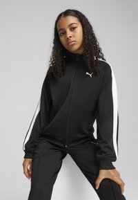 Crna trenirka s bijelim bočnim trakama, visokim ovratnikom, zatvaranjem na patentni zatvarač i Puma logom. Glatka textura, u kombinaciji s odgovarajućim crnim hlačama.