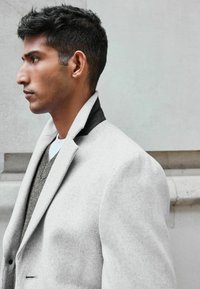 Abrigo de mezcla de lana gris claro con un collar negro, que presenta hombros estructurados y una textura suave, llevado sobre un suéter de punto gris.