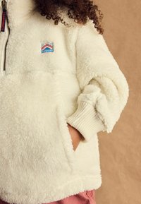 Veste en polaire blanche à col zippé quart, avec des poignets côtelés et une petite patch logo coloré sur la poitrine. Apparence douce et texturée.