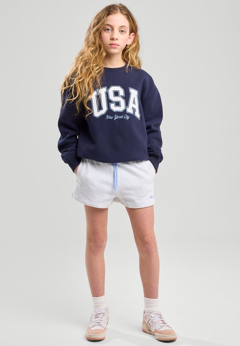 Sweatshirt marine avec "USA" et "New York City" imprimés en blanc. Associé à un short gris clair et des chaussettes blanches. Baskets avec des accents beige.