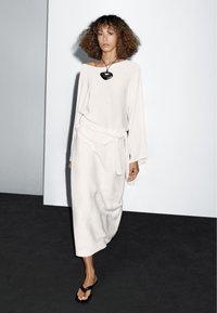 Donna con capelli ricci che cammina indossando un vestito bianco a maniche lunghe, una collana con pendente nero e sandali neri su un pavimento scuro con sfondo di parete chiara.