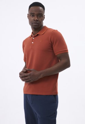 Mann steht mit gefalteten Händen, trägt ein rostfarbenes kurzärmeliges Poloshirt und dunkelblaue Hose vor schlichtem Hintergrund.