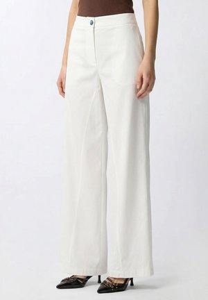 Pantaloni bianchi a vita alta e gamba larga con un bottone turchese, indossati con décolleté nere con punta affilata e una maglia marrone.