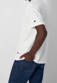 Camiseta de algodón blanca de manga corta, con un logo en el pecho y un parche en el lado. Combinada con unos jeans de mezclilla azul oscuro.