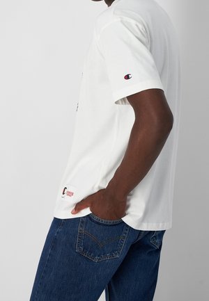 Hvid bomulds t-shirt med korte ærmer, prydet med et logo på brystet og et mærke på siden. Parret med mørkeblå denim jeans.