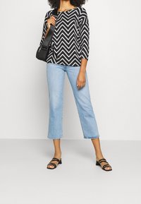 Schwarz-weißer Zickzack gemusterter Oberteil mit Dreiviertelärmeln, kombiniert mit hellblauen, taillierten Jeans und schwarzen, riemenförmigen, Absatzsandalen.