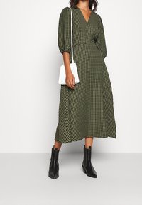 Robe à carreaux verts avec un décolleté en V, des manches bouffantes et une taille cintrée. Portée avec des bottes noires à la cheville et un sac crossbody blanc.
