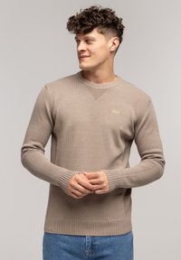 Beżowy sweter z dzianiny o okrągłym dekolcie, z prążkowanymi mankietami i dołem, z subtelną naszywką logo na klatce piersiowej, w połączeniu z niebieskimi dżinsami.