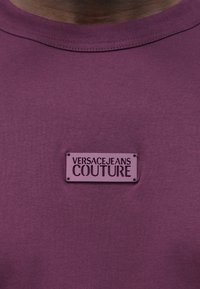 Pogled od blizu na ljubičastu majicu s kružnim izrezom s pravokutnom oznakom koja u sredini grudi glasi "VERSACE JEANS COUTURE".