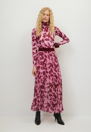 Woman with long curly blonde hair wearing a pink and burgundy floral long-sleeve dress and black ankle boots, standing against a plain background.