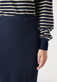 Pull rayé marine avec des rayures horizontales crème et une jupe navy côtelée. Présente un design ajusté et une texture douce, visible au niveau de la taille et des mains.