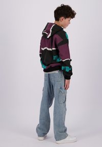 Flerfarget hettegenser i lilla, svart og teal med abstrakte mønstre. Løs passform cargo jeans i lyseblå med lommer og hvite sneakers.
