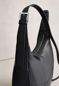 Sac à bandoulière en cuir noir texturé avec une sangle réglable et une boucle argentée, doté d'une fermeture éclair sur le dessus et d'un design minimaliste.