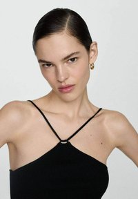 Črn rebrast halter top z tankimi naramnicami in osrednjim obročastim detajlom. Gladka tekstura z oprijetim krojem, nosi ga model s počešeno pričesko.