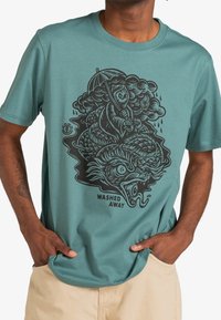 T-shirt de algodón de color azul verdoso con un diseño gráfico negro de un dragón y una figura sosteniendo un paraguas, con el texto "WASHED AWAY" debajo.