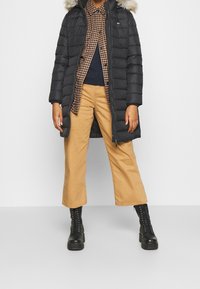 Abrigo acolchado negro con capucha forrada de piel, combinado con una camisa de cuadro de espiga marrón, una blusa azul marino, pantalones de pierna ancha beige y botas negras con cordones.
