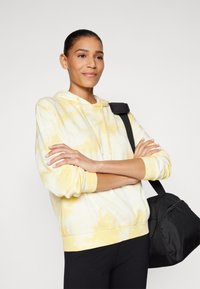 Haut à capuche tie-dye jaune et blanc avec un cordon de serrage, des poignets et un ourlet côtelés, porté avec des leggings noirs et un sac noir. Texture lisse.