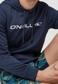 Mörkblå hoodie med dragsnören och vit "O'Neill"-logotyp, i kombination med mönstrade gröna och blå shorts med vågmönster.