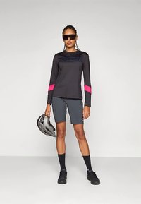 Černý cyklistický dres s dlouhým rukávem a růžovými detaily, doplněný šedými šortkami. Model drží šedou helmu, má na sobě tmavé sluneční brýle a černé boty.