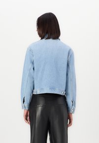 Giacca di jeans azzurra chiara con un design accorciato, dettagli con bottoni e fessure laterali, abbinata a pantaloni in pelle nera.