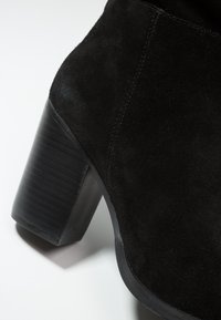 Bottine en daim noir avec un talon bloc épais et texturé et un design supérieur lisse et incurvé. Détail de couture latérale visible.