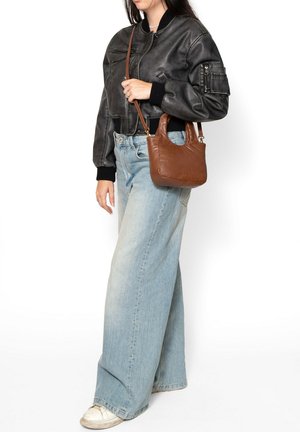 Femme portant une veste bombardier en cuir noire et un jean large bleu clair, avec un petit sac à main en cuir marron porté à l'épaule.