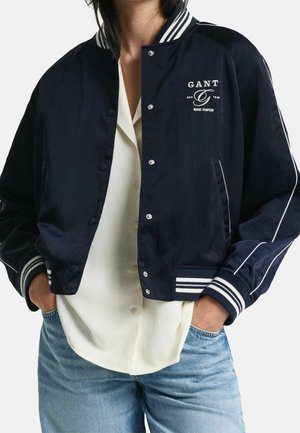 Osoba nosi granatową kurtkę varsity z białymi paskami, logo "GANT New Haven", białą koszulę na guziki oraz niebieskie dżinsy, ręce w kieszeniach.