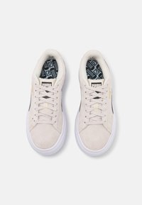 Paire de baskets Puma beige clair avec semelles blanches et lacets, bande latérale noire et semelles intérieures marquées, vues de dessus sur un fond gris.