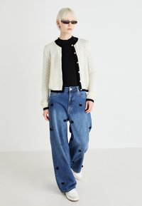 Cardigan bianco a maglia con rifiniture nere, top nero, jeans oversize blu con adornamenti floreali e sneakers bianche. Estetica minimalista.