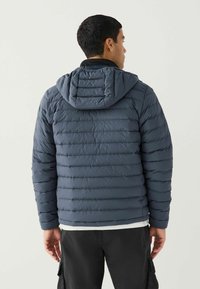 Tumši zilā puffer jaka ar horizontālu izšuvuma rakstu, regulējamu kapuci un elastīgām aprocēm. Tam ir rāvējslēdzēja aizdare un viegla materiāla sastāvs.