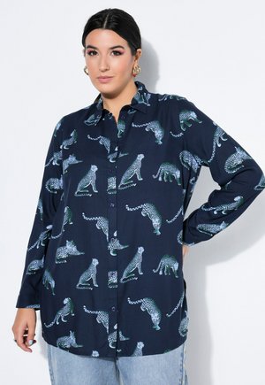 Kvinde iført en mørkeblå skjorte med knapper med flerfarvede gepardtryk og lyseblå jeans, stående med den ene hånd på hoften.