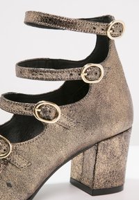 Chaussure à talon texturée métallique avec trois brides de cheville ajustables ornées de boucles ovales dorées.