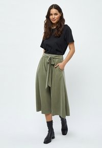 Must värviga lühikeste varrukatega T-särk, paari koos oliivroheline, kõrge vöökohaga culottes, millel on eesrihm. Mudel kannab musti pahkluuni ulatuvaid saapaid.