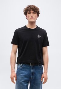 Νεαρός με σγουρά μαλλιά, φορώντας μαύρο t-shirt Calvin Klein και μπλε τζιν, στέκεται μπροστά από ένα απλό ανοιχτό φόντο.