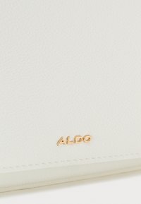 Materiale in pelle bianca con una finitura testurizzata. Presenta un logo "ALDO" dorato impresso sulla superficie. Bordo cucito in modo ordinato.