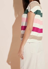 Kvinna med en kortärmad randig skjorta i beige, grönt, rosa och magenta, kombinerad med ljusa byxor mot en neutral bakgrund.