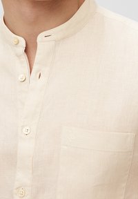 Camicia di lino beige con colletto alla mandarino, bottoni frontali e taschino sul petto con logo ricamato discreto.