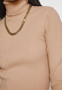 Pull à col roulé côtelé beige porté avec un collier épais en chaîne d'or, montrant le haut du torse et le cou.