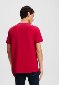 Tricou roșu din bumbac cu guler rotund, mâneci scurte și croială lejeră. Prezintă un logo mic aproape de tiv și o textură netedă.