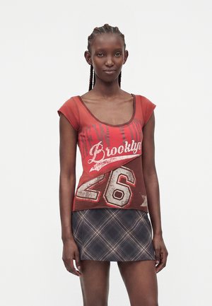 Ung kvinde med fletninger iført en rød grafisk t-shirt med teksten "Brooklyn 26" og et mørkt ternet mininederdel stående mod en hvid baggrund.