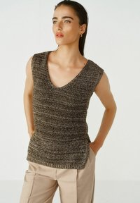 Top sans manches tricoté en marron foncé avec un col en V, présentant un motif texturé et une coupe ajustée. Associé à un pantalon beige.