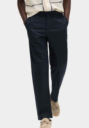 Persona che indossa pantaloni navy larghi con coulisse e tasche, abbinati a mocassini beige con nappine e una camicia chiara con ricami scuri.
