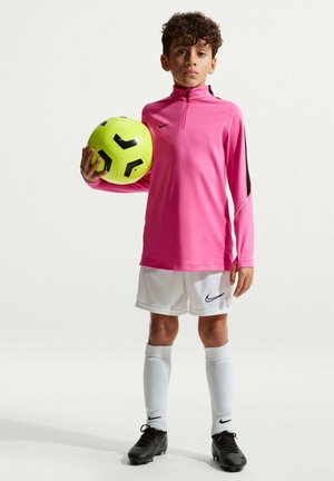 Dreng i pink langærmet sportstrøje, hvide shorts og sorte fodboldstøvler holder en gul og sort fodbold og står mod en ensfarvet baggrund.