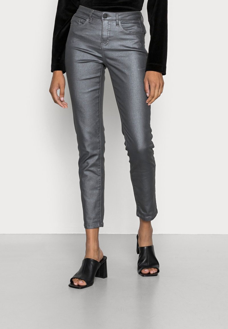 Femme portant un pantalon gris métallique ajusté et des mules à talons hauts noires à bout ouvert, debout devant un arrière-plan uni.