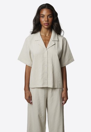 Kobieta w jasnobeżowej, teksturowanej, krótkim rękawem, zapinanej na guziki koszuli oraz pasujących szerokich spodniach na tle jednolitego białego tła.
