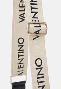 Canvas shoulder strap in beige, featuring black "VALENTINO" text. Silver hardware buckle connects two strap sections. Soft texture.