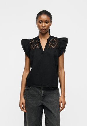 Kobieta w czarnej, bezrękawnikowej bluzce z falbankami na ramionach i koronkowymi zdobieniami na górnej części klatki piersiowej, w połączeniu z ciemnoszarymi dżinsami.