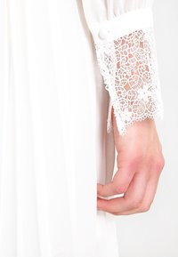 Abito bianco con una texture trasparente e maniche lunghe; rifinitura in pizzo sui polsini; chiusura con bottoni; tessuto morbido; design aderente.