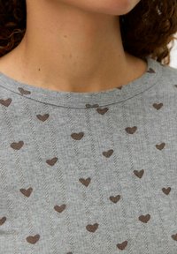 Grå kortärmad skjorta med rund halsringning, med en texturerad tyg och små bruna hjärtmönster överallt.
