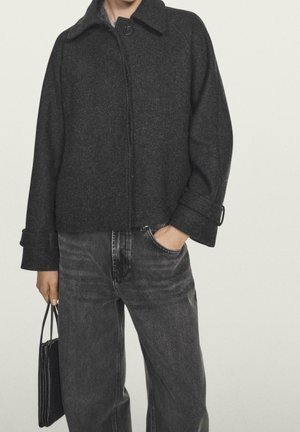 Person iført en mørkegrå uldjakke og falmede sorte jeans, der holder en lille sort håndtaske med den ene hånd i lommen.